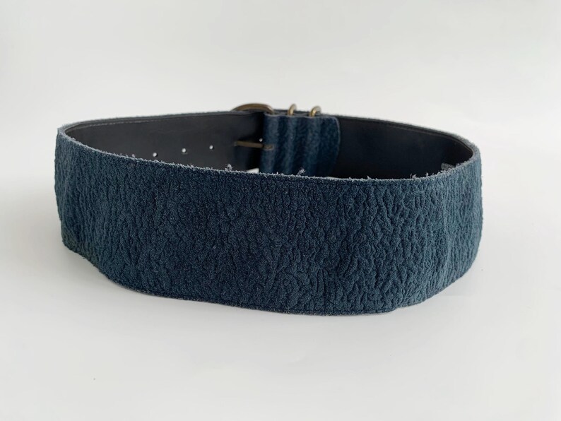 May include: A wide, navy blue belt with a textured finish. It features a metal buckle and adjustment holes. The belt is probably made of leather or a similar material.