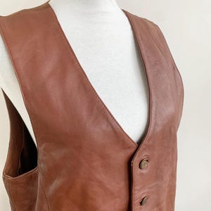 May include: A brown leather vest with a V-neck and three buttons. The vest is displayed on a white mannequin. The leather has a smooth texture and a warm, reddish-brown color.