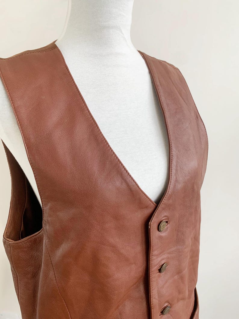 May include: A brown leather vest with a V-neck and three buttons. The vest is displayed on a white mannequin. The leather has a smooth texture and a warm, reddish-brown color.