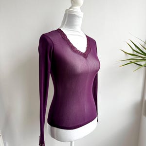 May include: A sheer, long-sleeved, plum-colored top with a V-neckline. The neckline and cuffs are trimmed with delicate lace. The top is displayed on a white mannequin.