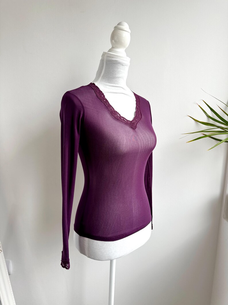 May include: A sheer, long-sleeved, plum-colored top with a V-neckline. The neckline and cuffs are trimmed with delicate lace. The top is displayed on a white mannequin.