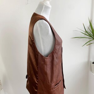 May include: A brown leather vest with a classic design. The vest features a V-neck, two front pockets, and a button closure. The vest is displayed on a mannequin.