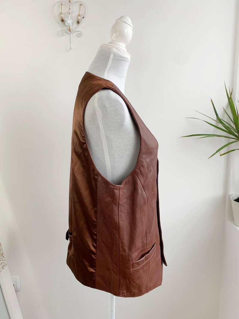May include: A brown leather vest with a classic design. The vest features a V-neck, two front pockets, and a button closure. The vest is displayed on a mannequin.