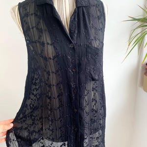 Puede incluir: Camisa negra sin mangas con cuello y botones. La tela transparente presenta un diseño floral bordado. La camisa se muestra en un maniquí.