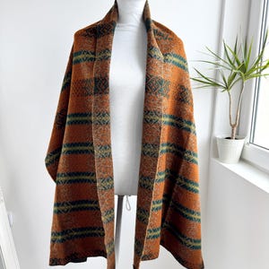Puede incluir: Un chal con estampado naranja y verde, colocado sobre un maniquí. El chal presenta rayas horizontales y diseños geométricos. La tela parece ser un material suave y tejido.