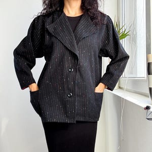 Puede incluir: Blazer negro con finas rayas verticales rojas, blancas y azules. El blazer tiene un cuello clásico, dos bolsillos delanteros y dos botones negros. Las mangas son largas y el forro es rojo.