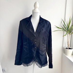 Pode incluir: Um blazer azul marinho escuro com gola entalhada e fecho de três botões. O blazer tem mangas compridas e uma bainha exclusiva com vieiras. A peça de roupa é exibida num manequim branco contra uma parede branca.