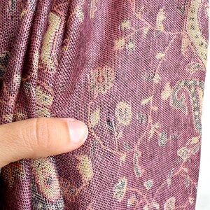May include: Close-up of a woven textile with a paisley pattern. The fabric is primarily a deep rose color with accents of beige, black, and light green. The design features floral and geometric elements, creating a textured appearance.