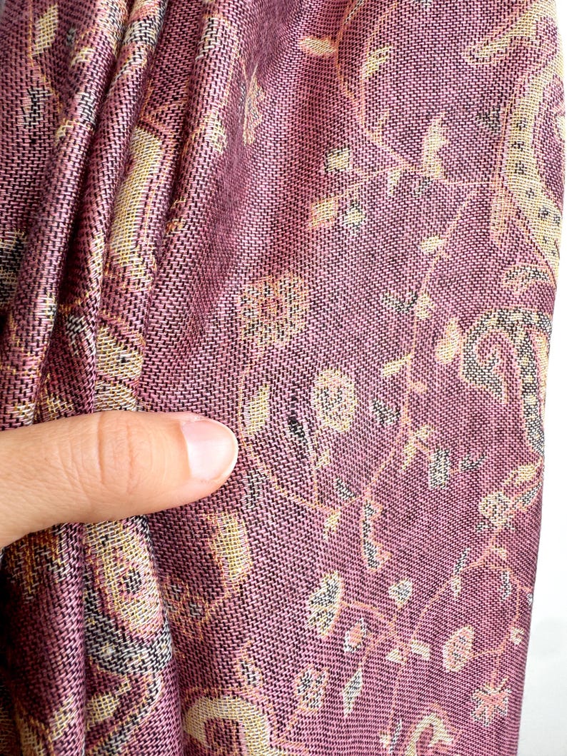 May include: Close-up of a woven textile with a paisley pattern. The fabric is primarily a deep rose color with accents of beige, black, and light green. The design features floral and geometric elements, creating a textured appearance.
