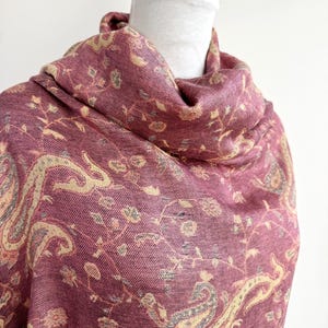 May include: A burgundy shawl with a detailed paisley and floral pattern. The scarf is draped over a white mannequin head. The design includes cream and gray accents, creating a textured and elegant look. The fabric appears soft and flowing.