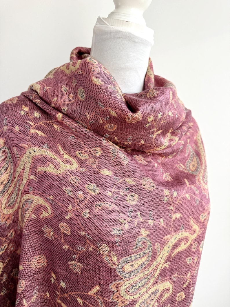 May include: A burgundy shawl with a detailed paisley and floral pattern. The scarf is draped over a white mannequin head. The design includes cream and gray accents, creating a textured and elegant look. The fabric appears soft and flowing.