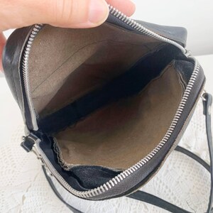 Pu&ograve; includere: Una borsa a tracolla in pelle nera con chiusura a zip e tiretto della zip argentato. La borsa &egrave; aperta e l'interno &egrave; foderato con un tessuto marrone.