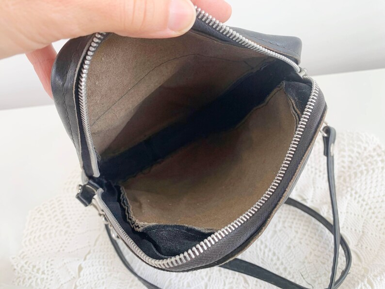 Pu&ograve; includere: Una borsa a tracolla in pelle nera con chiusura a zip e tiretto della zip argentato. La borsa &egrave; aperta e l'interno &egrave; foderato con un tessuto marrone.