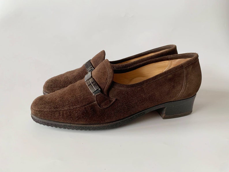 May include: Brown suede heeled loafers with a silver buckle detail.