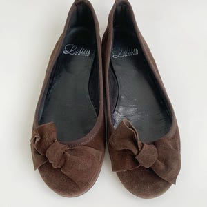 May include: A pair of brown suede ballet flats with a bow detail on the toe. The shoes have a rounded toe and a flat sole. The brand name "Lolita Milano" is printed on the insole.