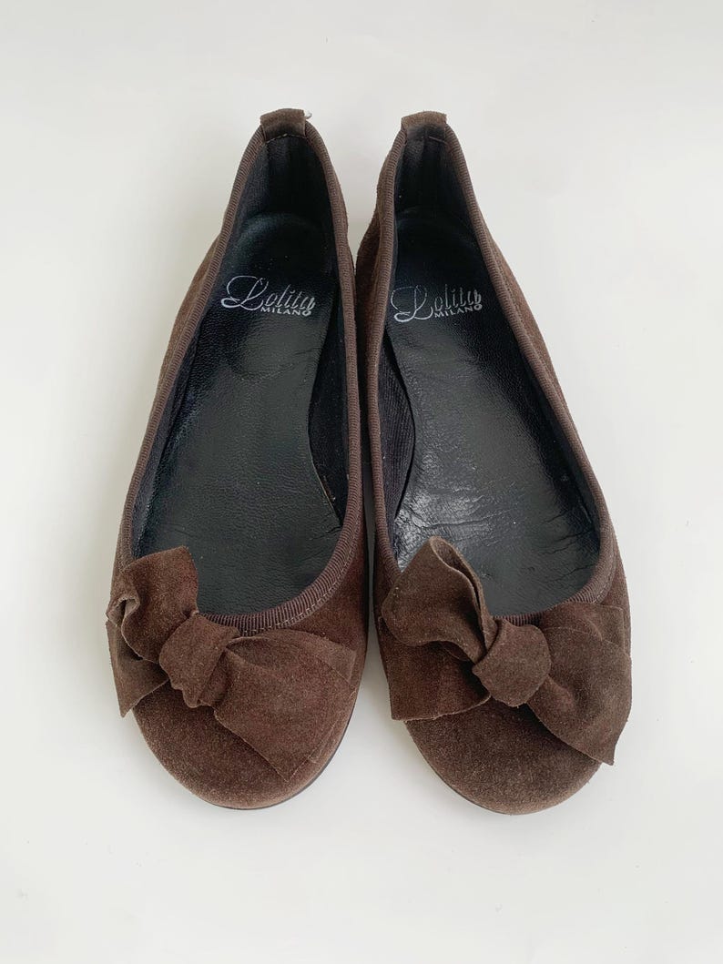 May include: A pair of brown suede ballet flats with a bow detail on the toe. The shoes have a rounded toe and a flat sole. The brand name "Lolita Milano" is printed on the insole.