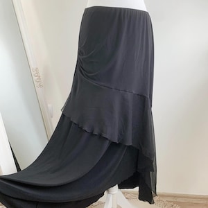 Puede incluir: Falda negra a capas con un diseño fluido. La falda presenta una cintura ajustada y un dobladillo asimétrico. La tela parece ligera, creando un efecto drapeado. La falda se exhibe en un maniquí.