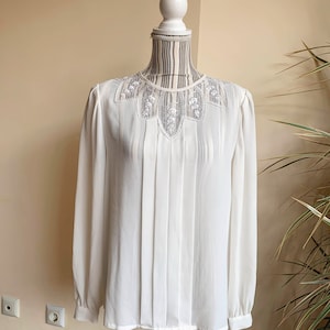 Puede incluir: Una blusa blanca de manga larga con un delicado cuello de encaje. La blusa tiene un frente plisado y un ajuste holgado.