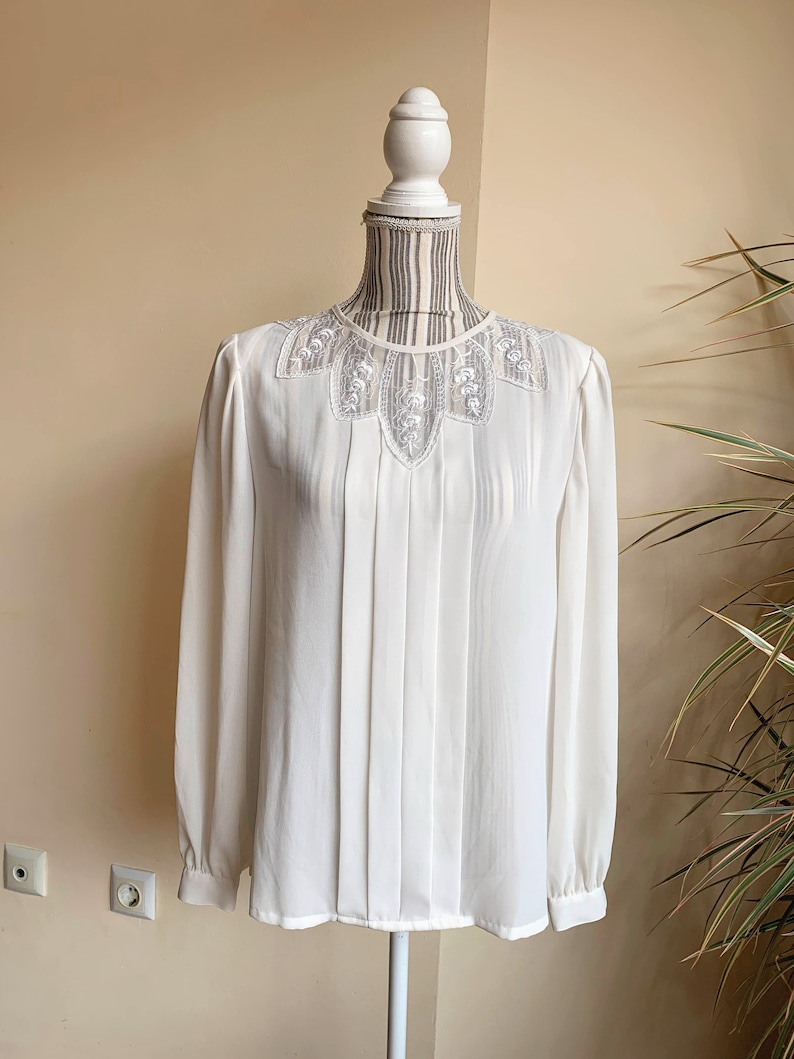 Puede incluir: Una blusa blanca de manga larga con un delicado cuello de encaje. La blusa tiene un frente plisado y un ajuste holgado.