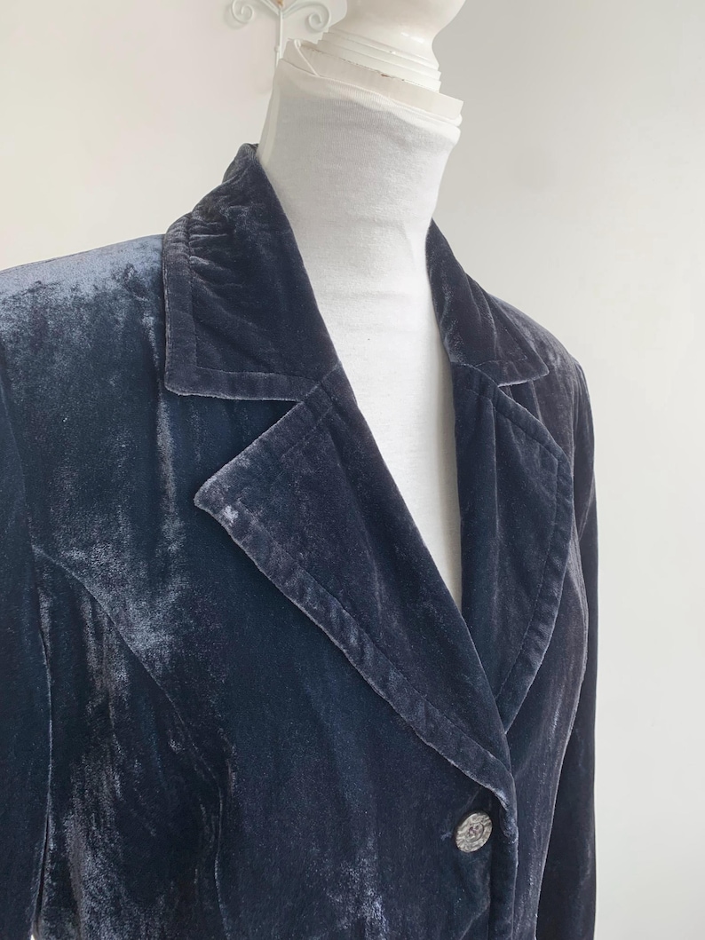 May include: A dark blue velvet blazer with a notched lapel and a single button closure. The blazer has a classic design and a soft, textured appearance. The button is silver with a decorative design.