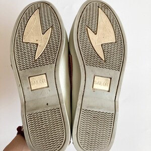 May include: A pair of light gray canvas shoes with brown leather accents. The soles are gray with a textured pattern and the word "SAILOR" is printed on each sole.