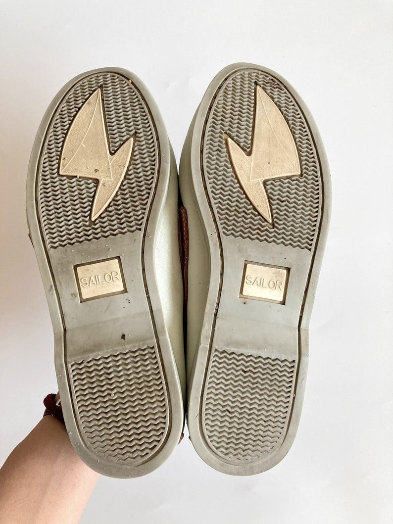 May include: A pair of light gray canvas shoes with brown leather accents. The soles are gray with a textured pattern and the word "SAILOR" is printed on each sole.