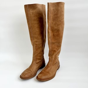 May include: A pair of knee-high brown leather boots. The boots have a rounded toe and a flat sole. The leather has a slightly distressed appearance. The boots are standing upright against a white background.