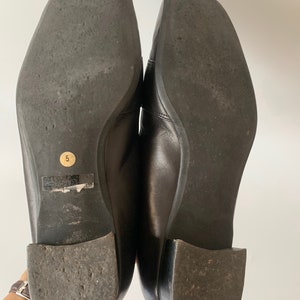 K&ouml;nnte beinhalten: Ein Paar schwarze Leder-Slipper mit quadratischer Spitze. Die Sohlen der Schuhe sind aus strukturiertem Gummi. Die Gr&ouml;&szlig;e 5 ist auf der Sohle eines Schuhs aufgedruckt.