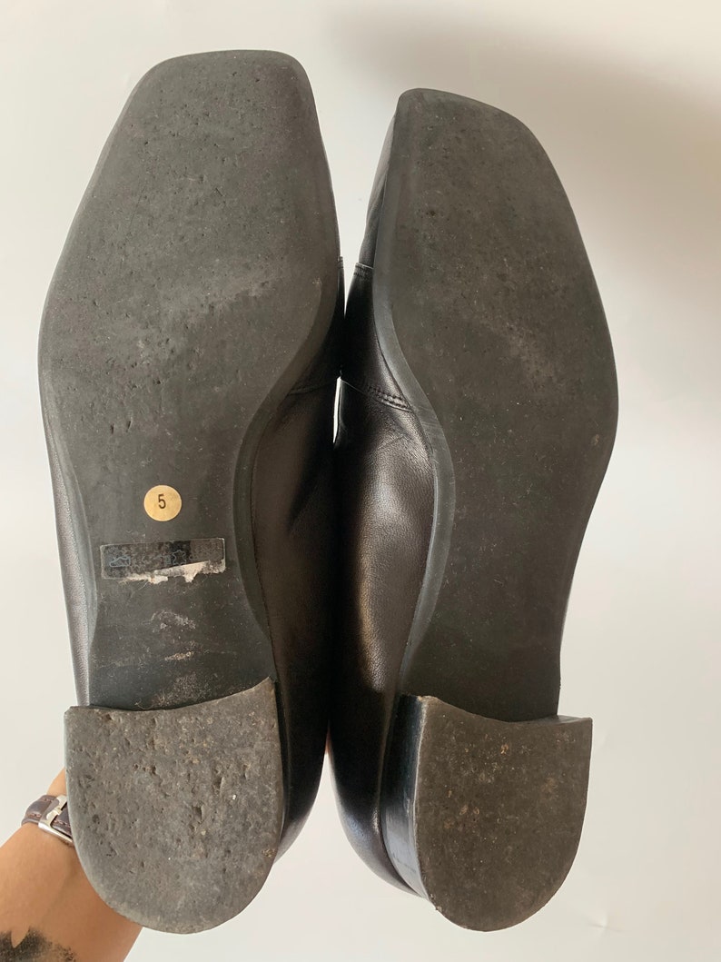 K&ouml;nnte beinhalten: Ein Paar schwarze Leder-Slipper mit quadratischer Spitze. Die Sohlen der Schuhe sind aus strukturiertem Gummi. Die Gr&ouml;&szlig;e 5 ist auf der Sohle eines Schuhs aufgedruckt.