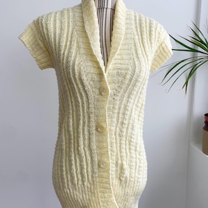 Puede incluir: Un cárdigan de punto amarillo pálido con cuello y cierre de botones. El cárdigan tiene mangas cortas y una textura acanalada.