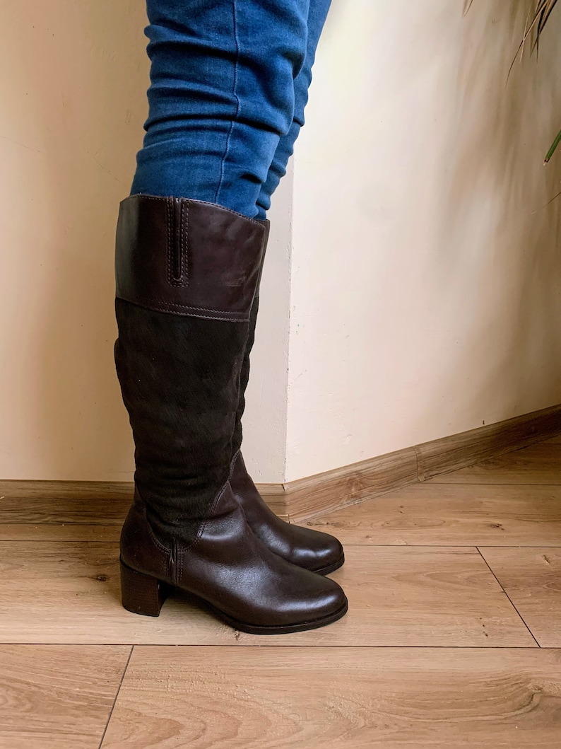 May include: Brown leather knee-high boots with a block heel. The boots have a suede panel on the back of the shaft.
