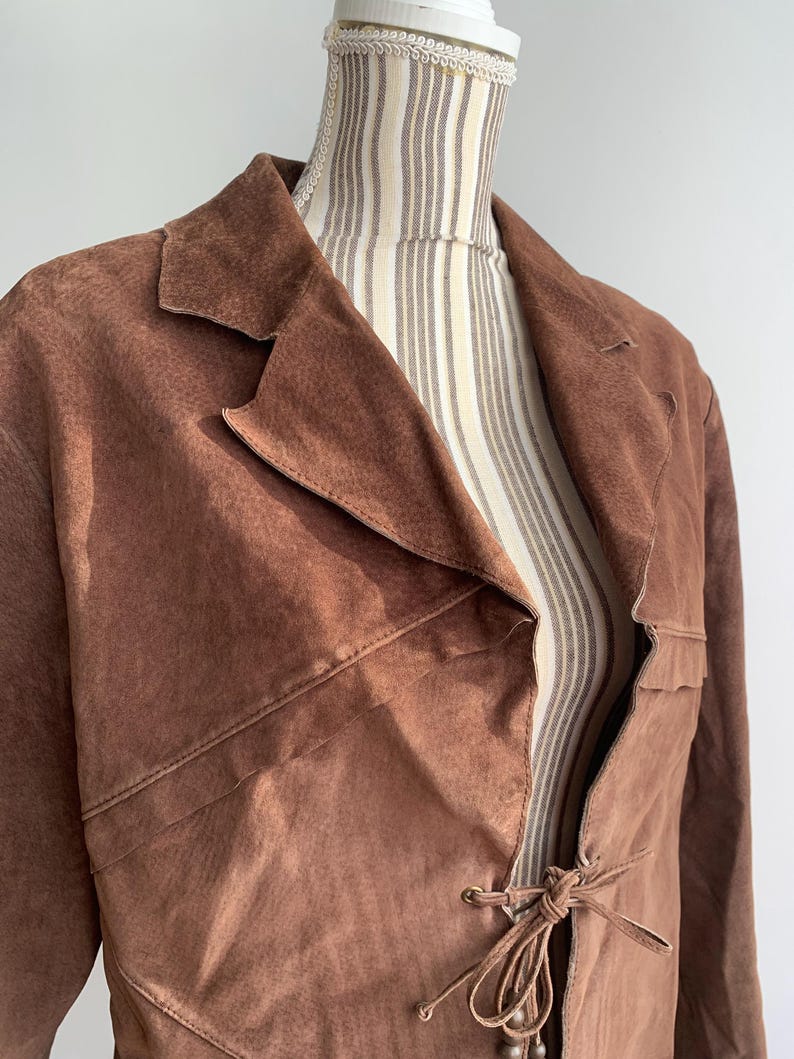 May include: A brown suede jacket with a long, flowing style. The jacket has a tie closure at the front and a collar.