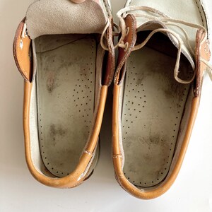 May include: A pair of white leather boat shoes with brown leather trim. The shoes have laces and are untied. The inside of the shoes is visible, showing a white leather lining with brown stitching.