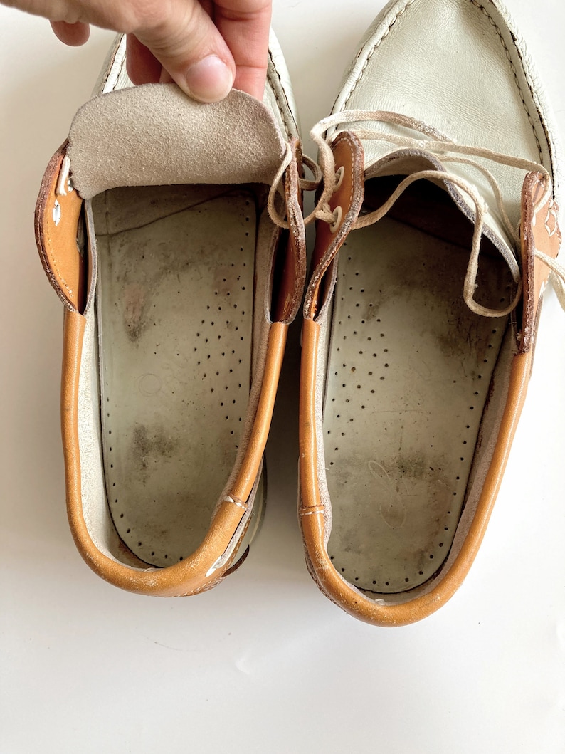 May include: A pair of white leather boat shoes with brown leather trim. The shoes have laces and are untied. The inside of the shoes is visible, showing a white leather lining with brown stitching.