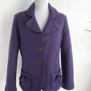 May include: A purple wool blazer with a notched collar, two front pockets, and three brown buttons. The blazer has a tailored fit and a textured appearance. The edges are trimmed with a darker purple. The jacket is displayed on a white mannequin.