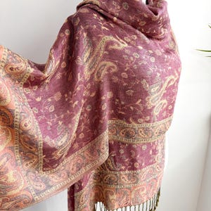 May include: A burgundy paisley patterned shawl with a fringed edge. The shawl features a detailed design in shades of gold, peach, and gray. The fabric is draped over a mannequin, showcasing the intricate pattern and texture of the textile.