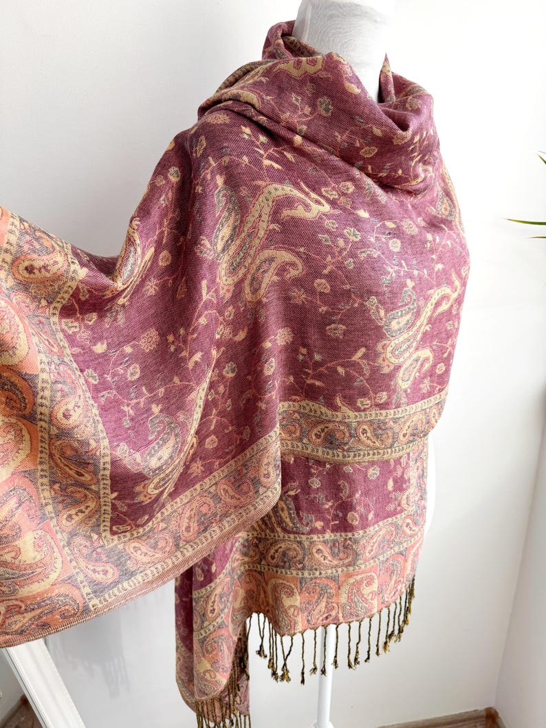 May include: A burgundy paisley patterned shawl with a fringed edge. The shawl features a detailed design in shades of gold, peach, and gray. The fabric is draped over a mannequin, showcasing the intricate pattern and texture of the textile.