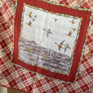 May include: A red and white plaid scarf with a white square in the center. The square features a landscape scene with ducks flying over a lake and a forest. The border of the square is a gold chain design.