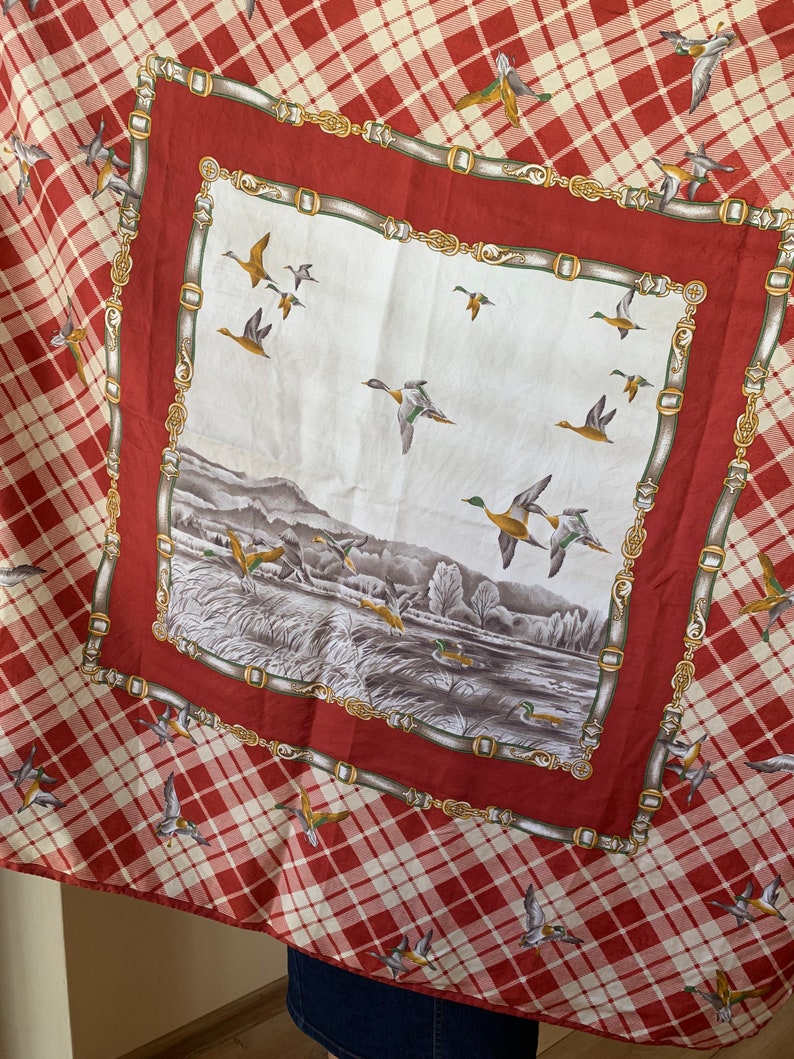 May include: A red and white plaid scarf with a white square in the center. The square features a landscape scene with ducks flying over a lake and a forest. The border of the square is a gold chain design.