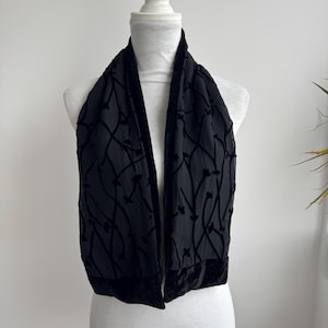 May include: A black scarf featuring a sheer, semi-transparent base fabric with a raised, velvet-like floral vine pattern. The scarf is finished with solid black velvet borders along the edges and the bottom hem, creating a textured contrast.
