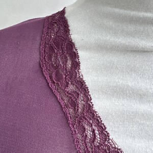 May include: Close-up of a plum-colored top with a V-neckline. The neckline is trimmed with a delicate, dark purple lace. The top is draped over a white surface, creating a contrast in color and texture.