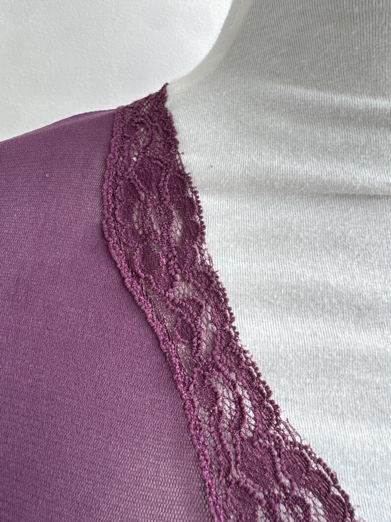 May include: Close-up of a plum-colored top with a V-neckline. The neckline is trimmed with a delicate, dark purple lace. The top is draped over a white surface, creating a contrast in color and texture.