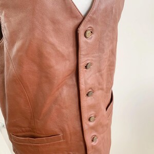 May include: A brown leather vest with a V-neck and a button-up front. The vest has two front pockets and is displayed on a white mannequin. The buttons are a brass color.