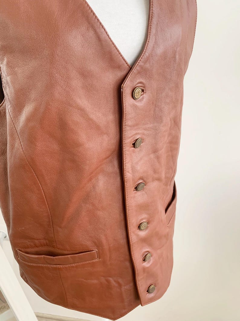 May include: A brown leather vest with a V-neck and a button-up front. The vest has two front pockets and is displayed on a white mannequin. The buttons are a brass color.