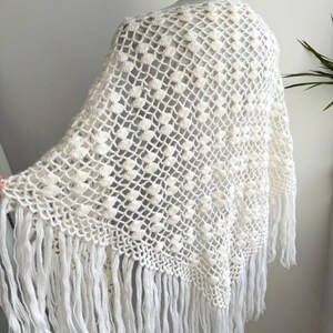 May include: Cream-colored crochet poncho with a diamond pattern and fringe. The open weave design features a textured, raised pattern. The long fringe hangs from the bottom edge. The poncho is displayed on a mannequin.