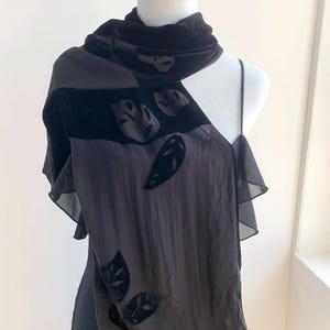 May include: A black scarf with leaf-shaped velvet appliqués is draped over a black dress. The scarf has a velvet collar and a sheer, flowing body. The dress has a simple, sleeveless design. The overall look is elegant and sophisticated.