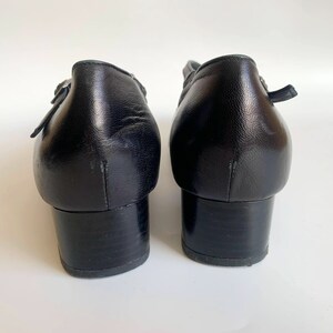 Pu&ograve; includere: Un paio di scarpe con tacco in pelle nera con cinturino con fibbia. Le scarpe hanno un tacco a blocco basso e una punta a punta.