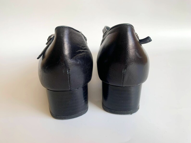 Pu&ograve; includere: Un paio di scarpe con tacco in pelle nera con cinturino con fibbia. Le scarpe hanno un tacco a blocco basso e una punta a punta.
