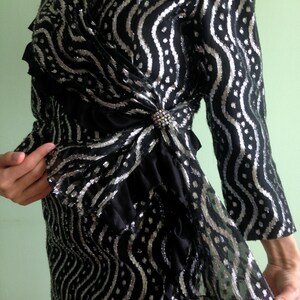 May include: A black and silver sequined dress with a wrap style bodice and a ruffled hem. The dress has a wavy pattern and is adorned with a silver brooch.
