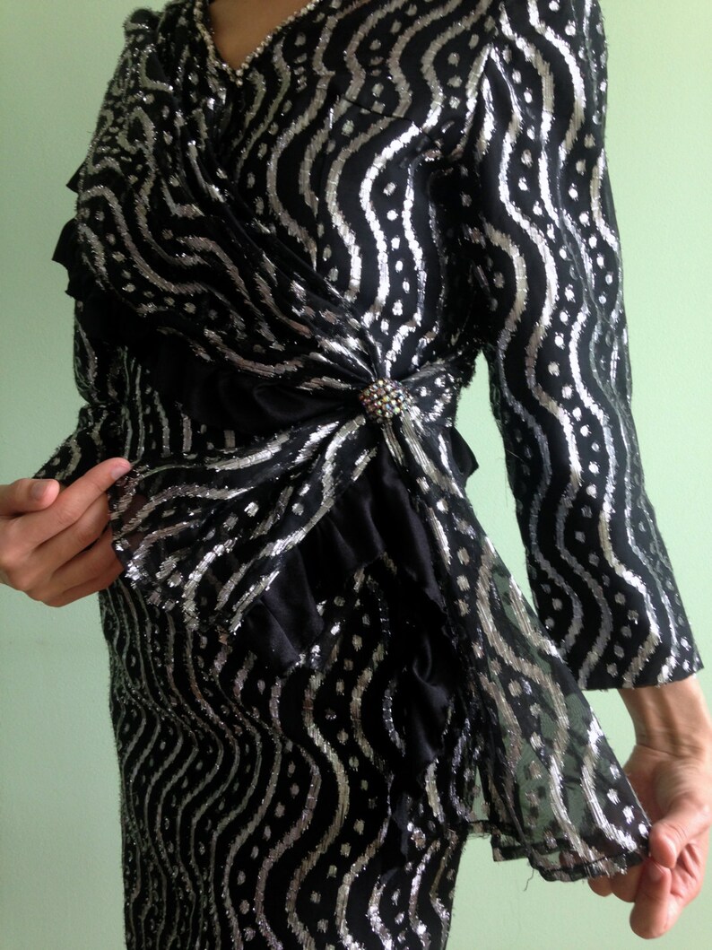 May include: A black and silver sequined dress with a wrap style bodice and a ruffled hem. The dress has a wavy pattern and is adorned with a silver brooch.